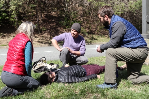 Three people outdoors kneeling around a fourth person who is lying on the ground.
