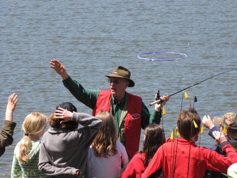 Volunteer Steve Sutter teaching a group of students how to fish. He has his right hand raised and holds a fishing pole in his left. Behind him is Hogback Pond with a purple hula hoop in it, used to help students learn to cast.