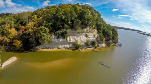 Tall white cliffs capped with overflowing vegetation jut out from the green and blue waters of a shimmering river. 