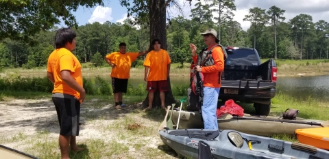 A group of people in orange shirts standing in a circle on a patch of sandy ground next to a black truck with two kayaks on the ground next to it