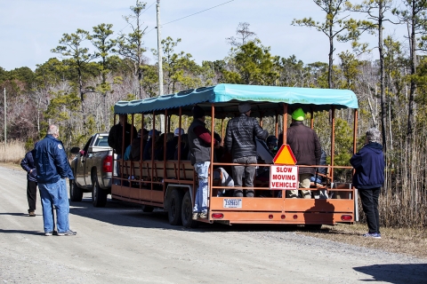 A large, open-air tram towed behind a pick-up truck pulled off on the side of the road, filled with passengers. A few volunteers stand to the side on the road.