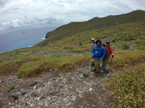 Eldridge Naboa, Ryan Peʻa, and Jiny Kim stand on a Nihoa. The ocean sits in the back. 