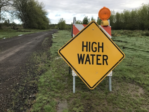 Bright yellow sign says "high water" to indicate high water levels on trail; sign is posted low to the ground next to gravel trail