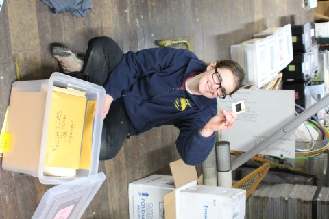 A young woman site cross-legged on a wooden floor, looking at a slide pulled from a tote full of files.