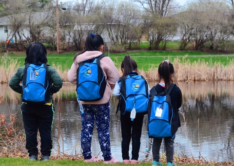Four kids face towards a pond with backpacks on