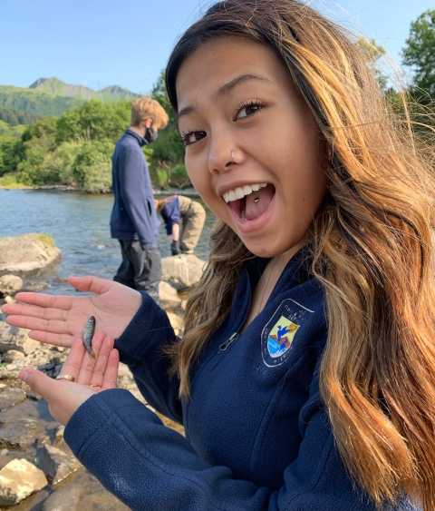 A smiling, enthusiastic young woman holding a very small fish next to a body of water