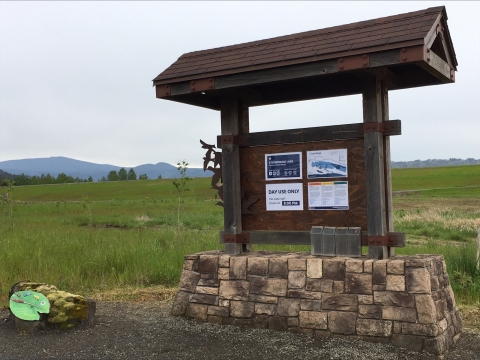 Information kiosk sits on top of a stone base with a small roof overhang to shield rain. Informational fliers are attached to the kiosk. A stone with an artists pallet sits a approximately three feet to the left. 