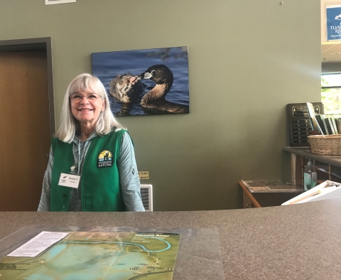 woman stands behind information desk; she is wearing a green vest with a logo and nametag. There is a laminated trail map on the counter in front of her, and a picture of waterfowl hanging on the wall behind her.