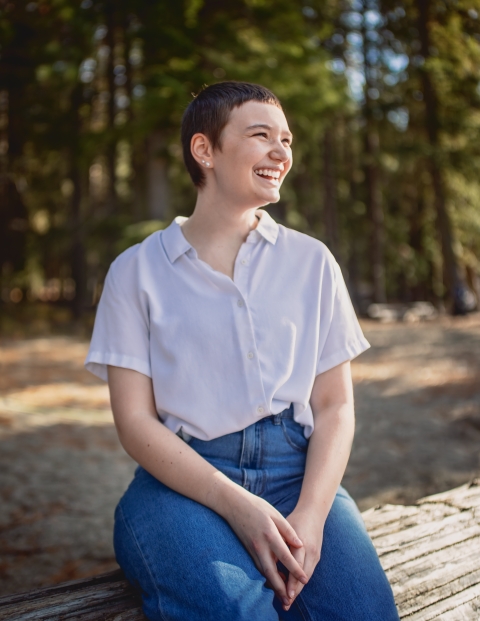 A smiling young woman in short sleeves and jeans sits on a log in a forested area.