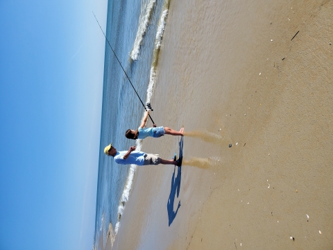 Fisherman teaching young girl to surf fish on the shore. The girl is holding a long fishing rod which has been cast into the surf. Small waves break on a sandy shore at the feet of the adult and child.