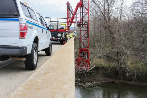 Person standing on a bridge over a river while a work truck extends a mobile catwalk beneath the bridge