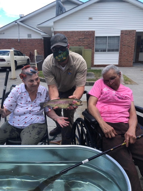 USFWS employee holding a Rainbow trout between two seated elderly women