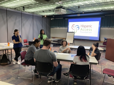 a group of people in a room sitting and standing around talking with a projector that reads "Hispanic access foundation" in the background.