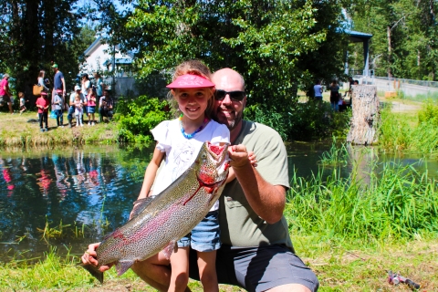 A dad in sunglasses holds a huge trout up in front of his daughter next to a fishing pond.