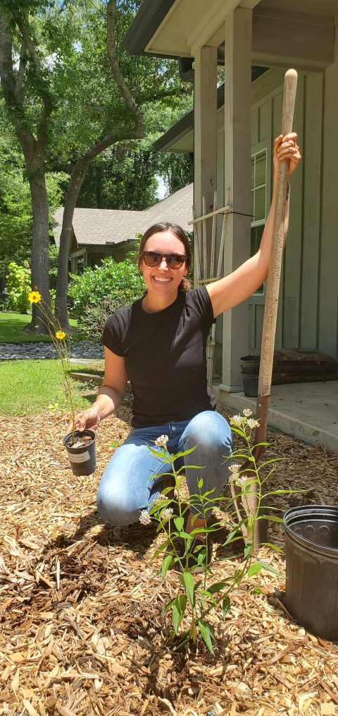 Jenna holding a shovel and a yellow flowered coreopsis plant posing for a picture before she plants the plant.   She is kneeling behind a milkweed plant.