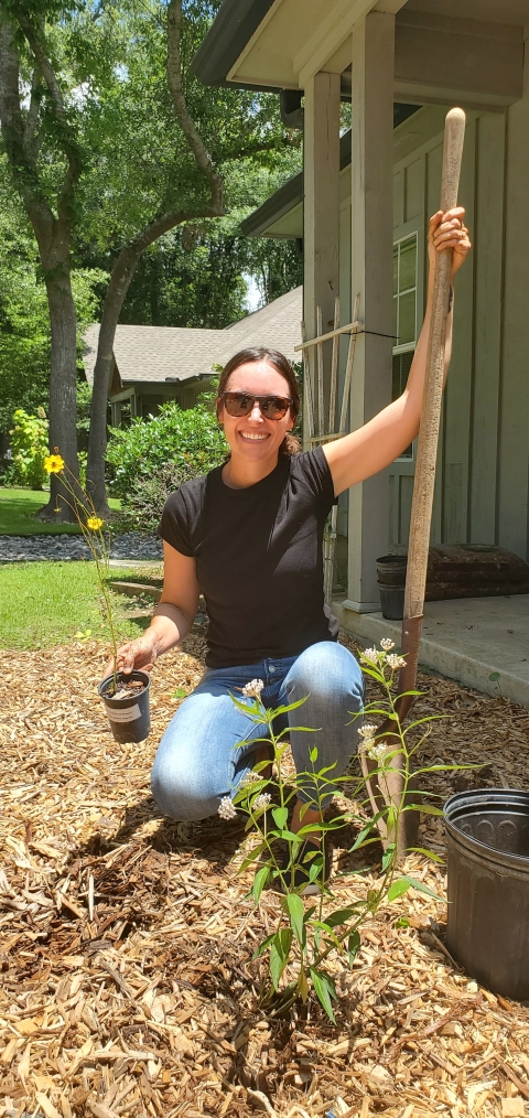 Jenna holding a shovel and a yellow flowered coreopsis plant posing for a picture before she plants the plant. She is kneeling behind a milkweed plant.
