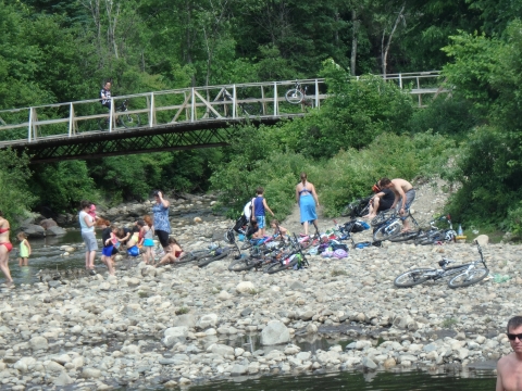 people recreate on a rocky riverside with a walking bridge in the distance