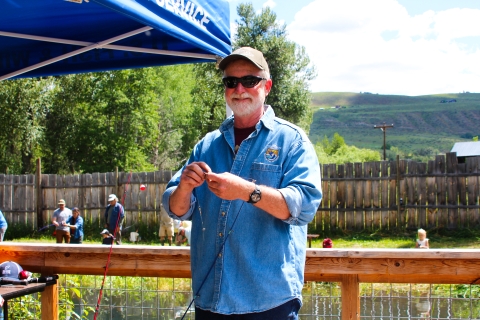 A man in a shirt with a Service logo, sunglasses, ballcap, white beard, and a smile prepares a fishing rod.