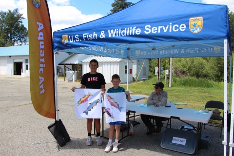 Two boys hold up bandanas with colored salmon on them, standing in front of a Service tent with a uniformed employee seated at a table and smiling.
