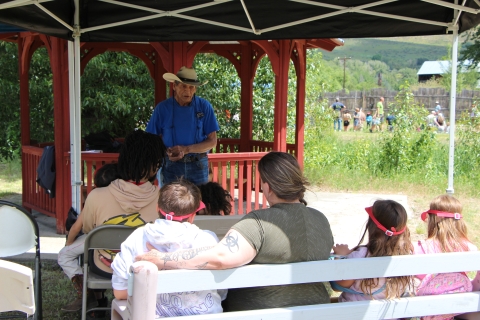 A man in braids and cowboy hat talks to a group of women and children seated in front of him.