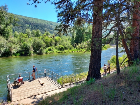 A man sits in a camp chair fishing from a platform with another man standing at the rail and two other people leaning on a rail beside the access trail, overlooking a river.