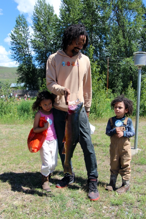 A smiling man holds a large rainbow trout for his two children to view.