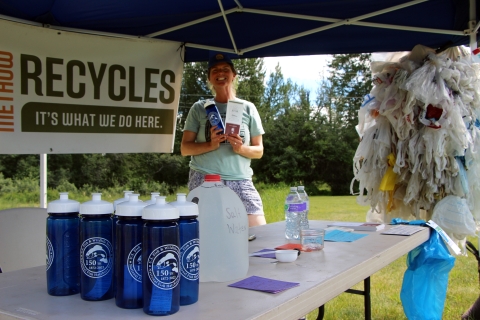 A woman in t-shirt and ballcap displays brochures while standing in a tent promoting recycling.
