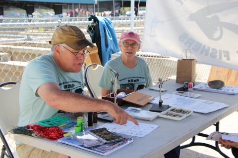 A man and woman in ballcaps and matching t-shirts sit at a table with fly-tying displays.