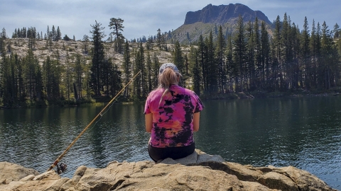 Woman with fishing pole sits on a rock overlooking the river with trees and mountains in the background