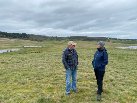 USFWS director, at right, talks with a local partner at a green pasture that is part of a western Oregon wildlife refuge, with gentle hills and water in the background.