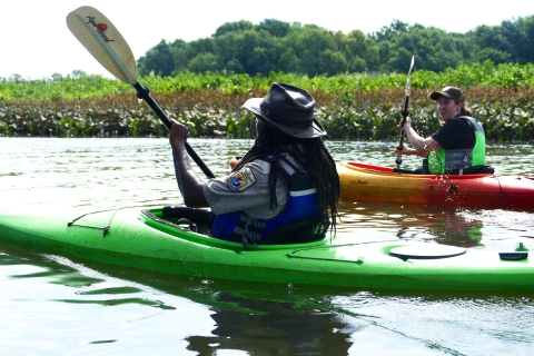 two Fish and Wildlife Service staff float side by side on the water in kayaks while holding oars. Marsh grass and trees can be seen in the background. 