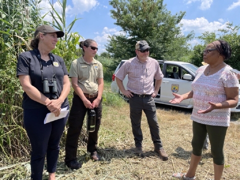 Four people stand together in a grassy marsh area speaking on a sunny day. A U.S. Fish and Wildlife van is parked behind them. 