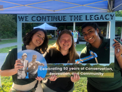 3 young women hold up a cardboard frame that says "We continue the legacy. Celebrating 50 years of conservation, recreation, and community
