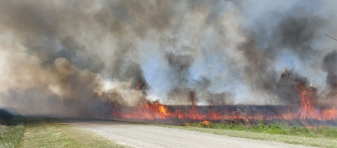 Fire on field with gravel road in between. Smoke fills the sky