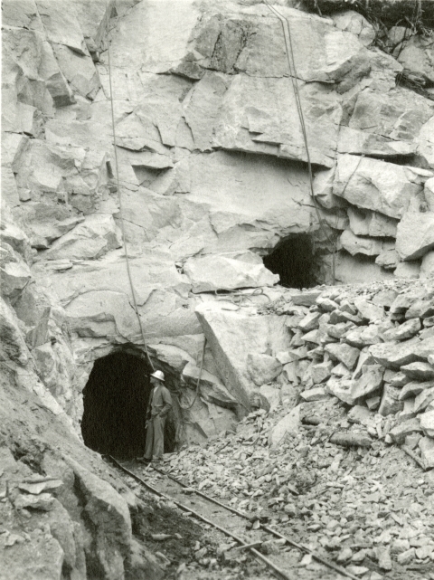 A black and white photo of a man in a hardhat standing at the lower entrance of two tunnels dug into a rock cliff with temporary rail tracks emerging.