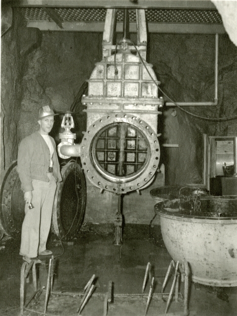 A black and white photo of a man in a hardhat standing beside a large piece of machinery in a cave hewn from rock.