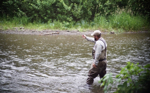 Man standing in river with water up to his knees, pointing upstream.