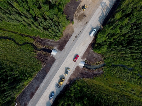 An aerial view of newly replaced culvert on a dirt road in rural Alaska. Cars and construction machines are parked on the road above the culvert, and a stream of water cuts through the greenery of pine trees and foliage.