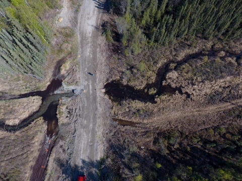 A small creek runs through an old and outdated culvert under a rough-looking rural dirt road in Alaska. The water is starting to erode the road due to not having a big enough opening to flow through.