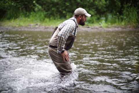 Man in light-colored clothing and fishing waders wades across a knee-high river.
