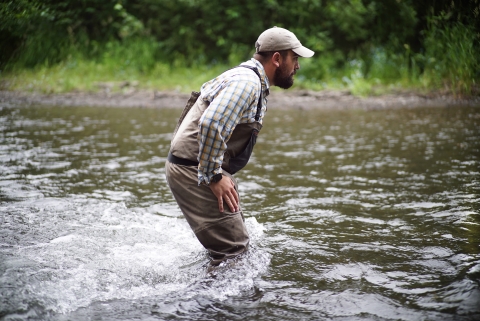 Man in light colored clothing, hat, and fishing waders wades across a knee-deep river.