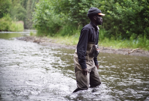 Man in dark clothing, hat, and fishing waders wades across a knee-deep river.