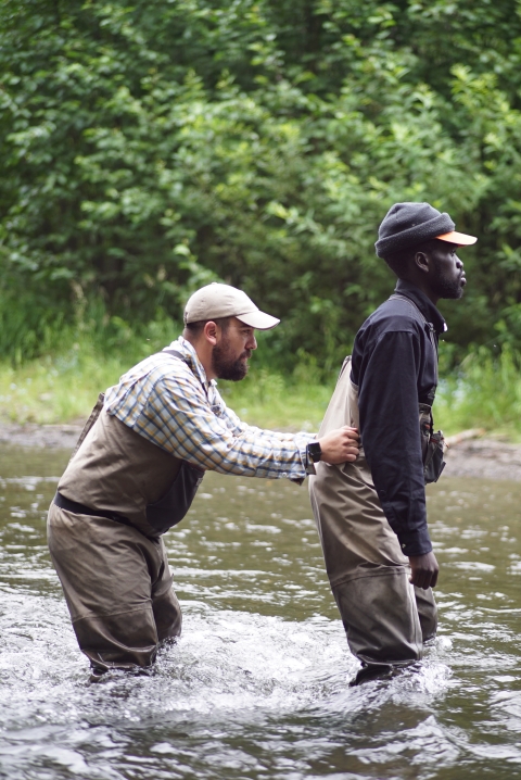 Two men are crossing a knee-deep river, and one is holding onto the other's waist to stabilize them while they cross.