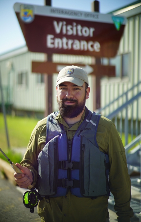 Man in hat and a green jacket wearing a personal floatation device (life jacket) holding a fishing rod.