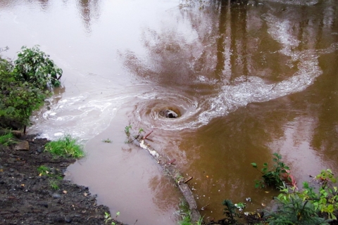 A whirlpool forms in the murky water above a culvert that is too small to handle the flow of water, and is being flooded.