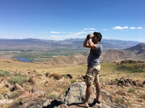 Man wearing t-shirt and cargo shorts stands at an angle while looking through a pair of binoculars. He stands on rocky terrain in a meadow and mountains in the far distance. 