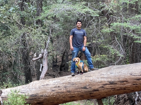 Man poses for a photo as he stands on downed tree trunk with dog sitting between his legs. 