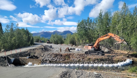 excavator digging near stream with mountains and clouds in background
