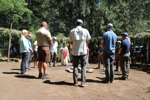 A group of people with heads slightly bowed stand in a circle in a forest