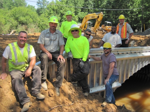 A group of people on a construction site pose next to a large culvert, smiling for the camera